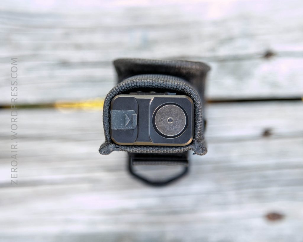 A close-up top view of a rectangular flashlight in a black fabric holster, resting on a weathered wooden surface. The flashlight’s two buttons and lens are visible.