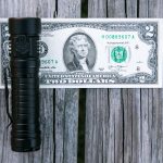 A black flashlight is placed vertically on top of a two-dollar bill, which is lying flat on a weathered wooden surface.