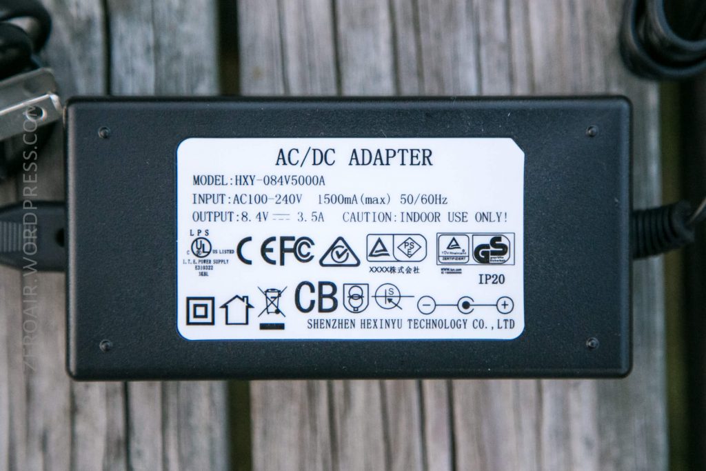 A rectangular black AC/DC adapter with a white label displaying technical specifications, safety symbols, and certification logos, resting on a wooden surface. The model number is HXY-084V5000A.