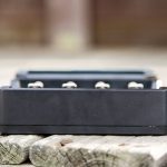 A close-up view of a black rectangular electronic device, possibly a battery charger, placed on a wooden surface outdoors. Four metal contacts are visible on the device. The background is blurred.