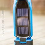 A compact blue device with two rectangular solar panels is standing upright on a wooden surface. The device has a black base and a clear cover over the solar panels.