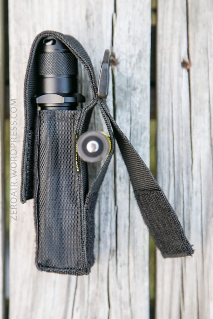 A black flashlight is secured inside a textured black carrying case with a hook-and-loop flap, resting on a weathered wooden surface. The side and front of the flashlight and case are visible.