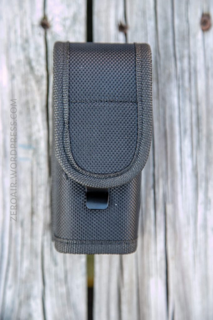 A black textured nylon pouch with a flap and snap closure is shown standing upright on a weathered wooden surface. The words zeroair.wordpress.com are faintly visible on the left side.