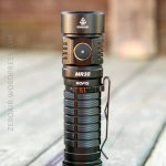 A black Rofis MR30 flashlight stands upright on a wooden surface. The flashlight has a metal clip and a prominent power button on its side. The background is blurred.