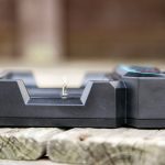 A black battery charger sits on a wooden surface outdoors. The charger is empty and viewed from the side, with a metal contact visible in the center. The background is blurred.