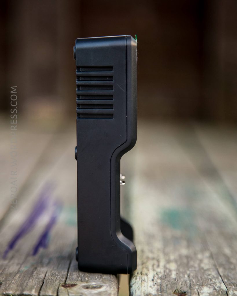 A black plastic electronic device stands upright on a weathered wooden surface, shown from the side. The background is blurred, drawing attention to the devices ribbed and smooth textures.