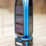 A blue portable lantern with built-in solar panels stands upright on a wooden surface. The device has a black base with a power button and visible solar cells on its sides.
