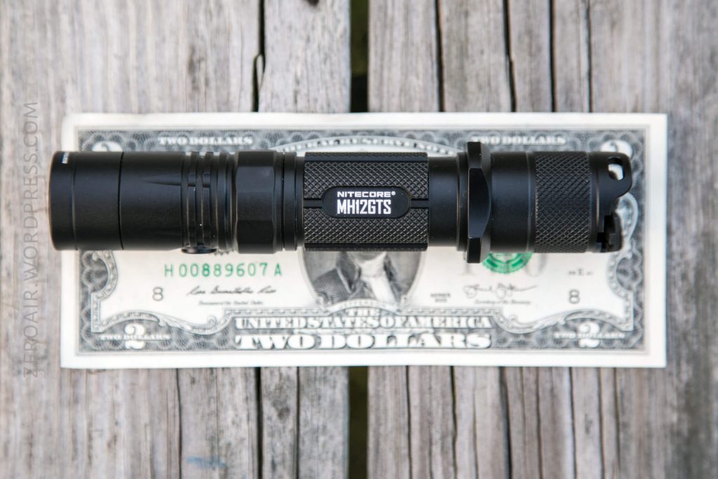 A black Nitecore MH12GTS flashlight is placed on top of a two-dollar bill, which is lying on a weathered wooden surface. The flashlight is roughly the same length as the bill.