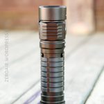 A black, cylindrical flashlight stands upright on a wooden surface, with a blurred background. The flashlight has a textured grip and ridged sections on the body.