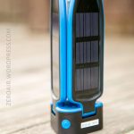 A blue solar-powered lantern stands on a wooden surface. The lantern features two black solar panels and a small blue power button on the base. The background is blurred.