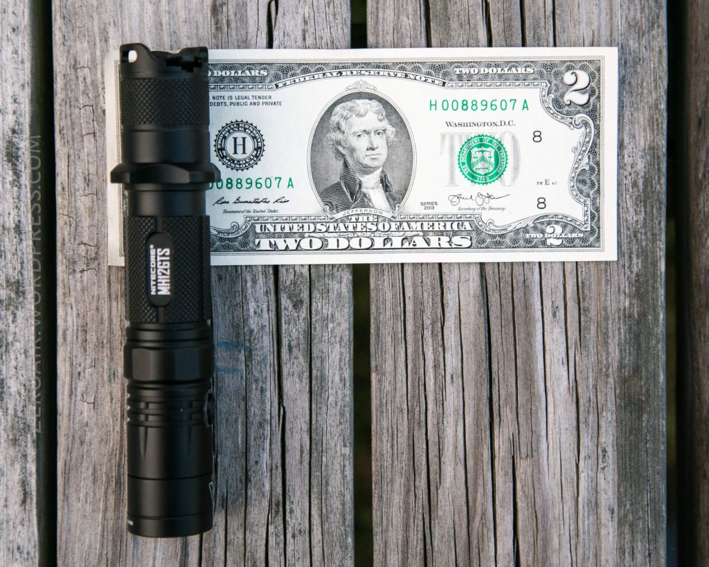 A black flashlight is placed next to a U.S. two-dollar bill on a wooden surface. The flashlight is slightly shorter than the width of the bill and both objects are viewed from above.