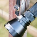 A black Thrunite flashlight is attached to a black strap with a metal clip, hanging from a wooden post outdoors. The flashlight brand name is visible on the side.