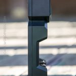 A black electronic device with an angular design is standing upright on a wooden surface, photographed from the side. The background is blurred.