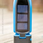 A compact blue device with a black base and a rectangular solar panel on the front is standing upright on a wooden surface. The background is blurred.