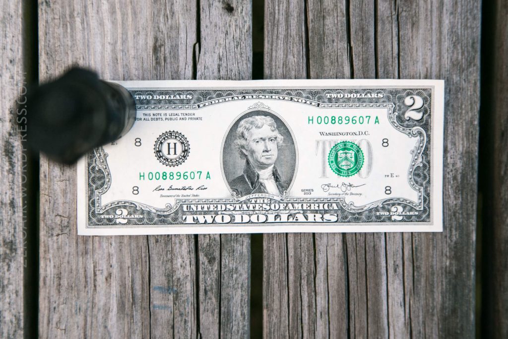 A U.S. two-dollar bill featuring Thomas Jefferson is placed on a wooden surface and held down by a black metal object in the upper left corner.