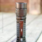 A metallic flashlight with a textured grip and a pocket clip stands upright on a weathered wooden surface. The background is blurred and brown-toned.