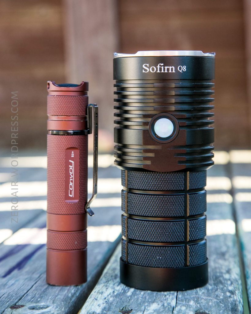 Two flashlights stand side by side on a wooden surface. The left flashlight is red and slim with a pocket clip labeled Convoy, while the right one is wider, black, and labeled Sofirn Q8 with a large central button.