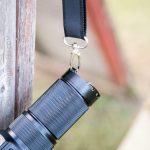 A close-up of a black flashlight attached to a lanyard by a metal keyring, hanging from the corner of a wooden surface. The background is blurred, showing an outdoor setting.