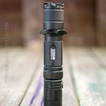 A black Nitecore MH27S flashlight stands upright on a weathered wooden surface with a blurred brown background. The flashlight has a textured grip and visible branding on the handle.