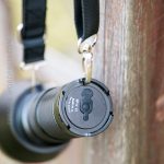 A close-up of a camera lens attached to a strap, hanging from a hook on a wooden fence. The lens cap has an inscription, and part of the strap hardware is visible.