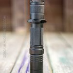 A black tactical flashlight stands upright on a weathered wooden surface, shown in clear focus against a blurred brown background.
