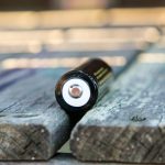 A close-up of a single cylindrical battery lying on its side on a weathered wooden surface, with the positive terminal facing the camera.