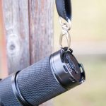 A close-up of a black textured flashlight hanging by a keyring from a strap, attached to a weathered wooden post. The background is blurred and out of focus.
