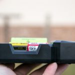 A close-up of a hand holding a black battery charging station with several batteries inserted. The background is blurred, and there is a watermark reading ZEROAIR.WORDPRESS.COM along the left side.