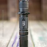 A black handheld flashlight stands upright on a weathered wooden surface outdoors. The flashlight has a textured grip and a label with a logo and the text “Rofis.” The background is out of focus.