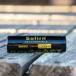 A Sofirn rechargeable 18650 lithium-ion battery with a capacity of 2200mAh is placed on a weathered wooden surface, shown in close-up with sunlight illuminating the scene.