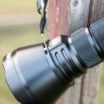 Close-up of a black handheld flashlight attached by a strap to a metal hook on a wooden post, showing textured grip and a power button. The word THRUNITE is partially visible on the flashlight.