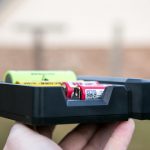 A hand holds a black battery charger with two cylindrical rechargeable batteries, one green and one red, inserted in the slots. The background is blurred.
