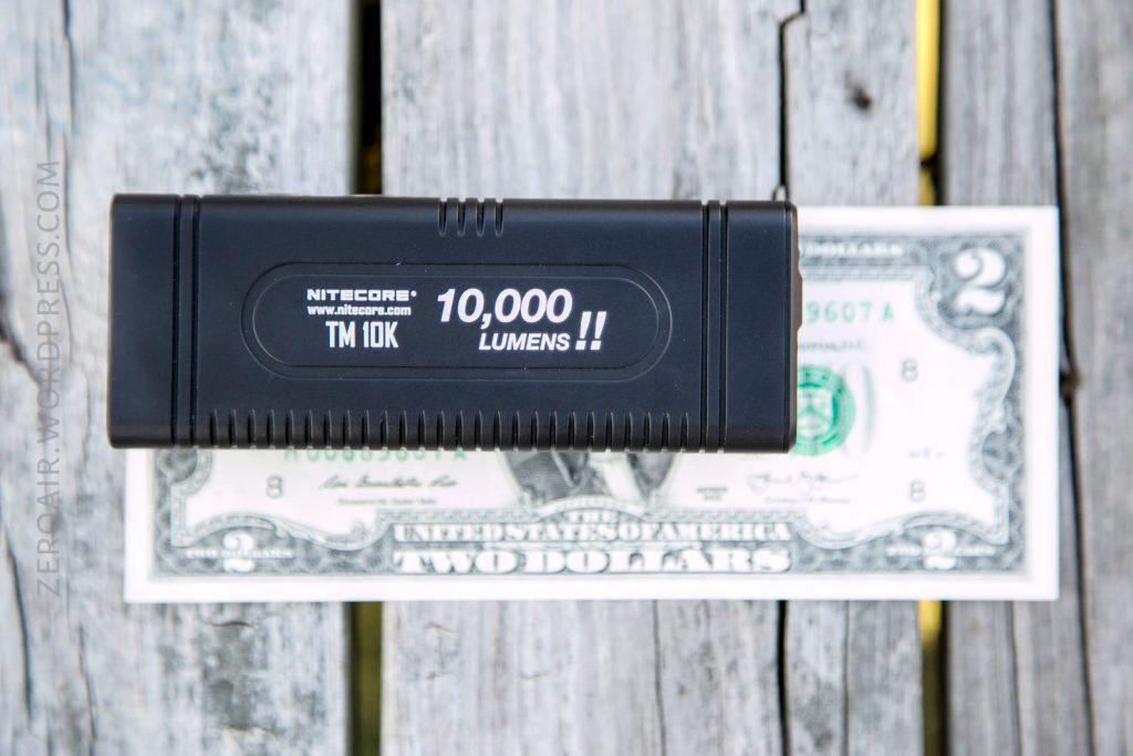 A black Nitecore TM10K flashlight labeled 10,000 lumens is placed on top of a two-dollar bill on a weathered wooden surface. The flashlight is shorter than the length of the bill.