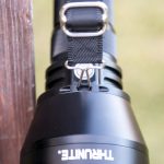 A close-up of a black THRUNITE flashlight with a black strap attached by a metal clip, resting against a wooden surface with a blurred outdoor background.