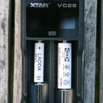A black XTAR VC2S battery charger holds two white IKEA LADDA AAA rechargeable batteries. The charger is placed on a weathered wooden surface.