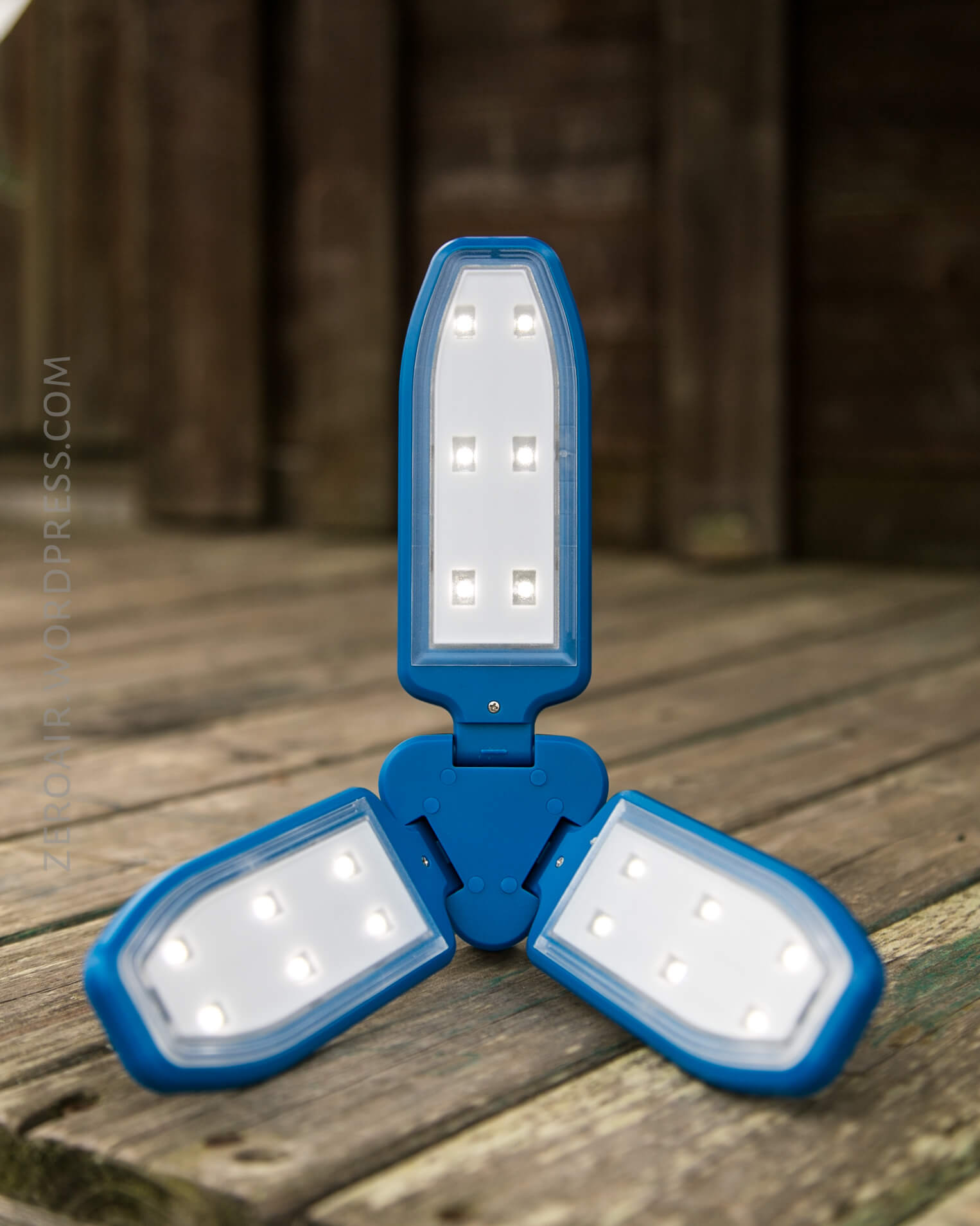 A blue, three-panel LED camping lantern is open and standing on a wooden surface outdoors. Each panel has lit LED lights, and the background is out of focus.
