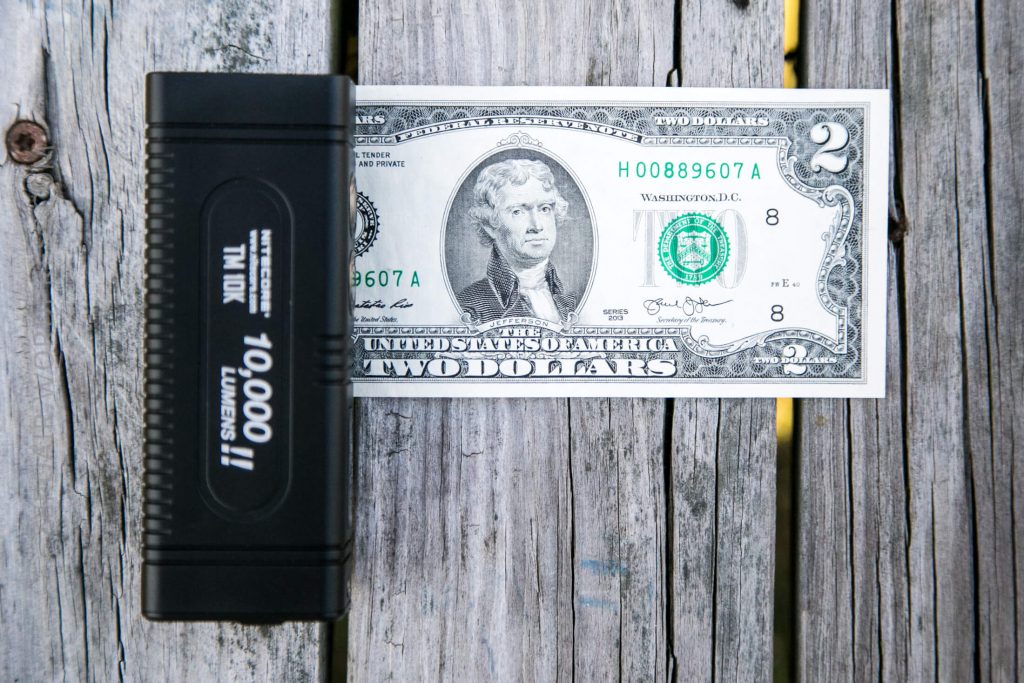 A black flashlight labeled 10,000 II LUMENS sits next to a U.S. two dollar bill on a weathered wooden surface. The bill features Thomas Jefferson’s portrait.