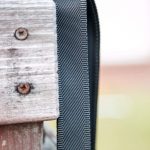 A close-up of a black nylon strap looped over a weathered wooden post with two rusted screws, set against a blurred outdoor background.