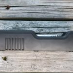 A black plastic rectangular object with ridges on one side sits on a weathered wooden surface, partially obscuring a cylindrical metallic item inside the slot.