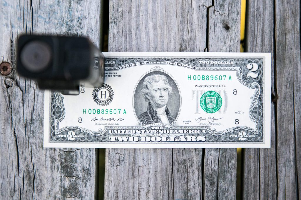 A black handgun is pointed at a two-dollar bill lying on a weathered wooden surface. The gun is in the upper left of the image, and the bill features Thomas Jefferson’s portrait in the center.