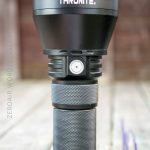 A black ThruNite flashlight stands upright on a wooden surface outdoors. The flashlight has a textured grip and a single button on its body. ZER0AIR.WORDPRESS.COM is faintly visible on the left side.