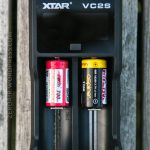 A black XTAR VC2S battery charger holding two cylindrical batteries, one red and one yellow, is placed on a weathered wooden surface. The brand name and model are visible on the charger.