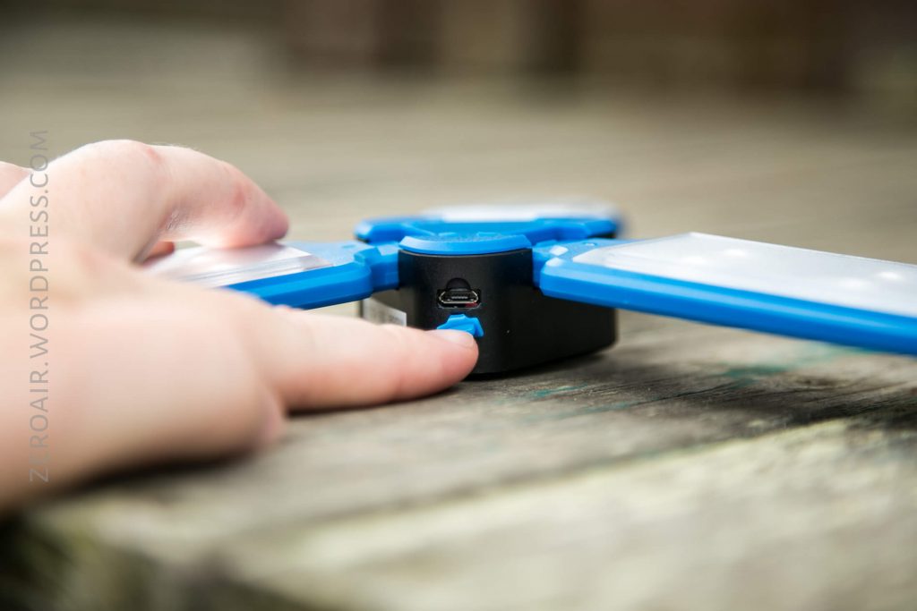 A close-up of a person’s hand pressing a button on a small, blue electronic device with foldable panels, resting on a wooden surface. A micro USB port is visible on the device.