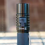 A close-up of a black flashlight with a textured blue grip, a pocket clip, and a visible USB charging port cover. Certifications and symbols are printed below the port. Background is blurred.