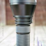 A large black handheld flashlight with a textured grip and wide head is standing upright on a wooden surface. The background is blurred with brown and green tones. The flashlight is the main focus.