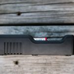 A black rectangular electronic device with vent-like grooves sits on a weathered wooden surface. Part of a small red and white object is visible inside a slot on the top edge of the device.