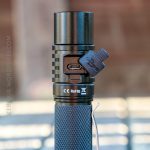 A close-up of a black flashlight with a textured grip, showing a USB-C charging port covered by a rubber flap. The flashlight has certification markings and is standing upright on a blurred surface.