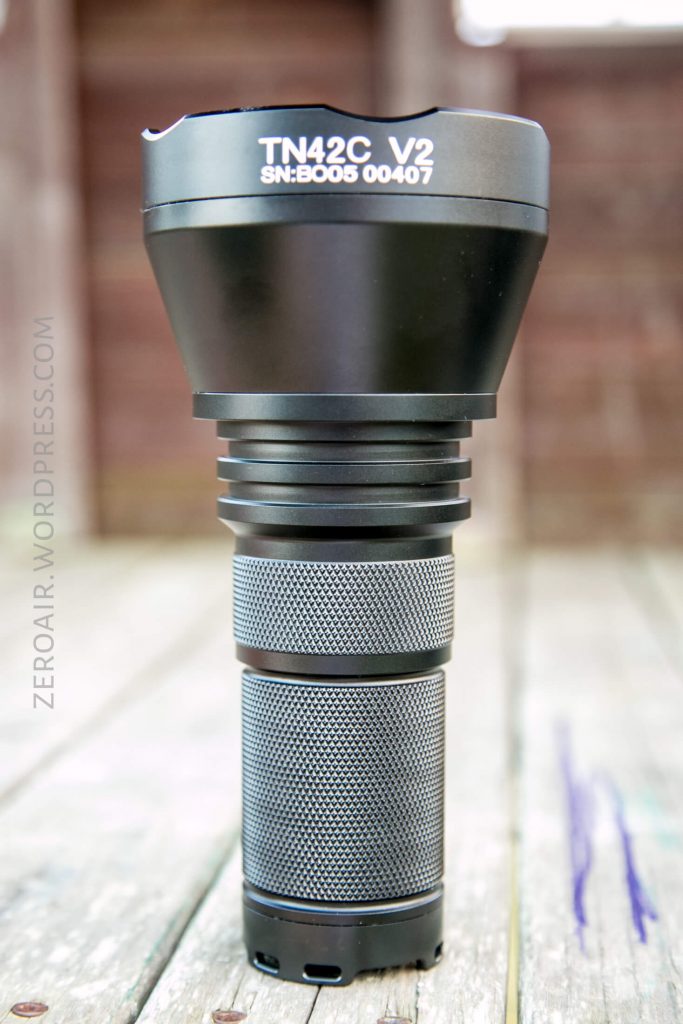 A black TN42C V2 flashlight with serial number B005 00407 is standing upright on a light-colored wooden surface. The flashlight has a wide head and a textured grip handle.