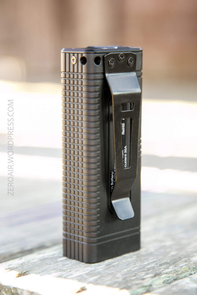 A black, rectangular flashlight with a textured surface, metal clip, and visible screws is standing upright on a wooden surface. The side of the device is marked with some text.