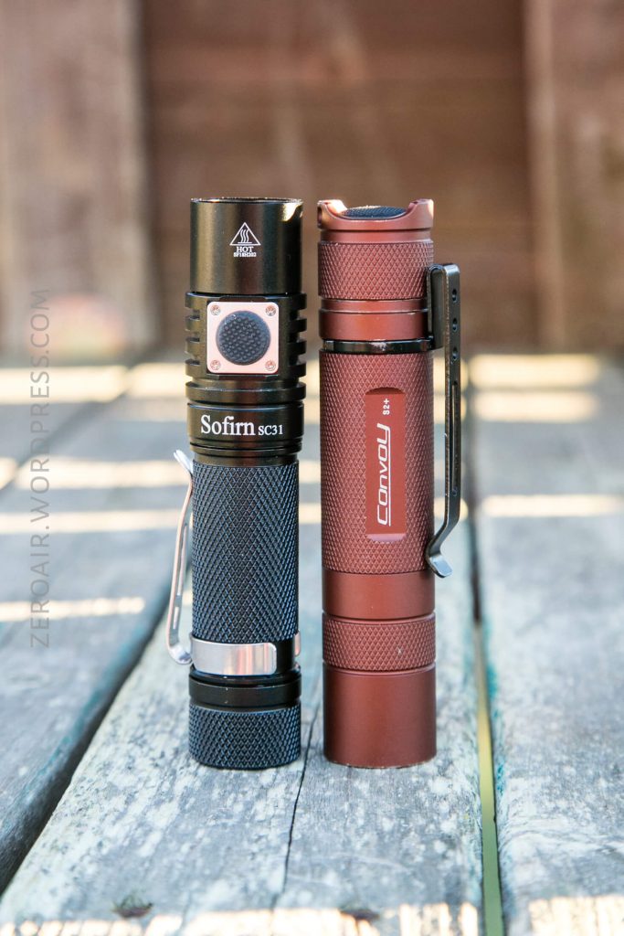 Two flashlights, one black Sofirn SC31 and one red Convoy S2+, standing upright side by side on a weathered wooden surface with a blurred brown background.